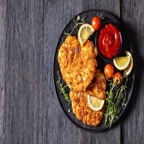 fried juicy chicken breast cutlets under crusting of panko breadcrumbs on black plate with tomato sauce, lemon, fresh thyme and ripe tomatoes on dark wood table, flat lay, free space