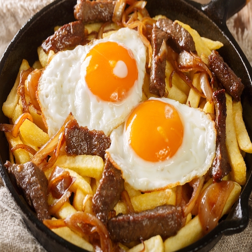 Chorrillana is a dish consisting of french fries topped with sliced meat, fried eggs, and onions close up in the pan on the table. Horizontal