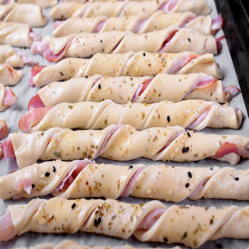 Sticks of raw puff dough with bacon sprinkled with sesame seeds on the oven tray ready to be baked