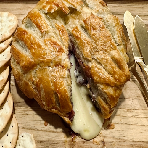 Baked brie, made with blueberry preserves, pictured with crackers on the side and cheese spreading knives.